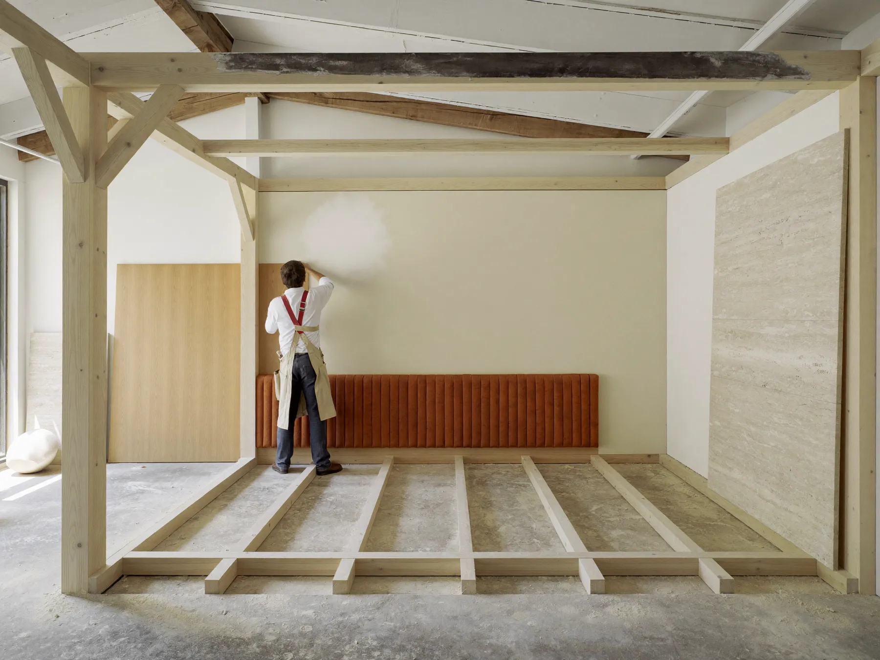Begehbarer Hotelzimmer-Prototyp im Rohbau mit Holzrahmen, verputzter Wand und terrakottafarbenem Polsterpaneel in einer hellen Werkhalle